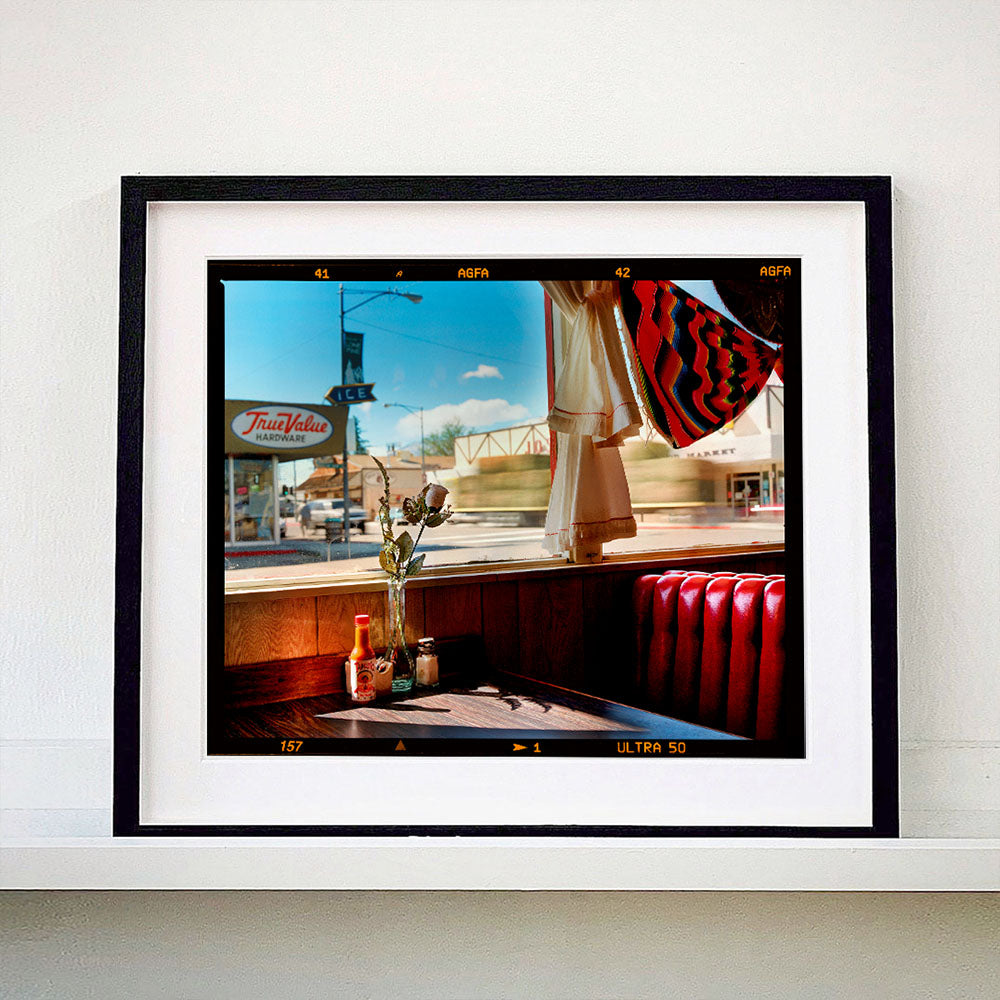 Black framed photograph by Richard Heeps. Inside an American diner the light is shining on the table and the seat is the classic vibrant red faux-leather of a retro diner. The photograph is looking out of the window into the outside street.