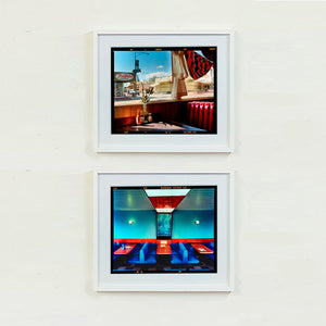 Two white framed photographs by Richard Heeps. The top one shows the Inside an American diner the light is shining on the table and the seat is the classic vibrant red faux-leather of a retro diner. The photograph is looking out of the window into the outside street. The bottom photograph is the inside of a Vintage Wimpy in Norfolk, bright blue retro seating and red tables, set against a blue wall.