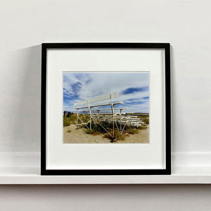 Black framed photograph by Richard Heeps. The back view of a basic white painted grandstand sits in the middle of this photograph. Grass grows up from its base and it sits alone on a sandy ground.