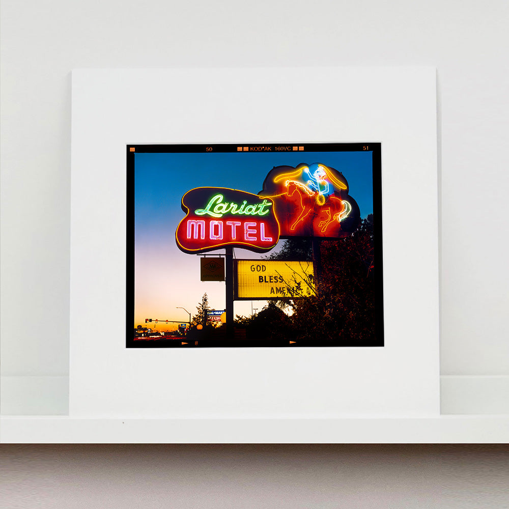 Mounted photograph by Richard Heeps. The photograph is taken in the evening, a neon sign belonging to the Lariat Motel, with a cowboy on horseback with a lasso on the right of the words, half way down the sign post is the yellow sign God Bless America.