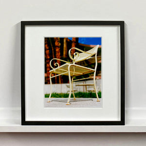 Black framed photograph by Richard Heeps. A cream chair sits on hard standing, behind the chair and slightly out of focus is lush green grass and warm red tree trunks.