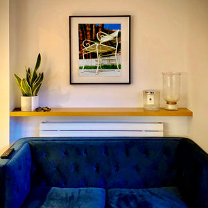 Black framed photograph in situ by Richard Heeps. A cream chair sits on hard standing, behind the chair and slightly out of focus is lush green grass and warm red tree trunks.