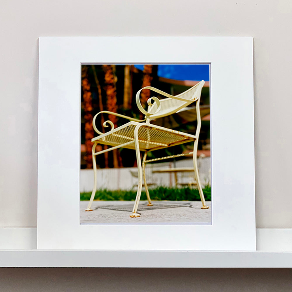 Mounted photograph by Richard Heeps. A cream chair sits on hard standing, behind the chair and slightly out of focus is lush green grass and warm red tree trunks.