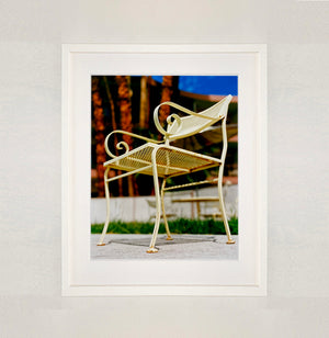 White framed photograph by Richard Heeps. A cream chair sits on hard standing, behind the chair and slightly out of focus is lush green grass and warm red tree trunks.