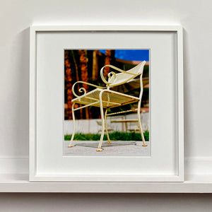 White framed photograph by Richard Heeps. A cream chair sits on hard standing, behind the chair and slightly out of focus is lush green grass and warm red tree trunks.