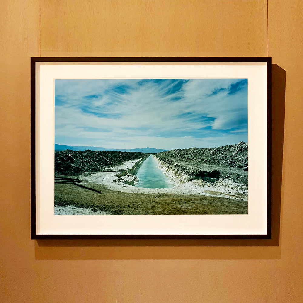 Black framed photograph by Richard Heeps. A watery white trench is dug into the centre going into the distance with grey mounds of earth either side. The sky is vast and blue, with light clouds.