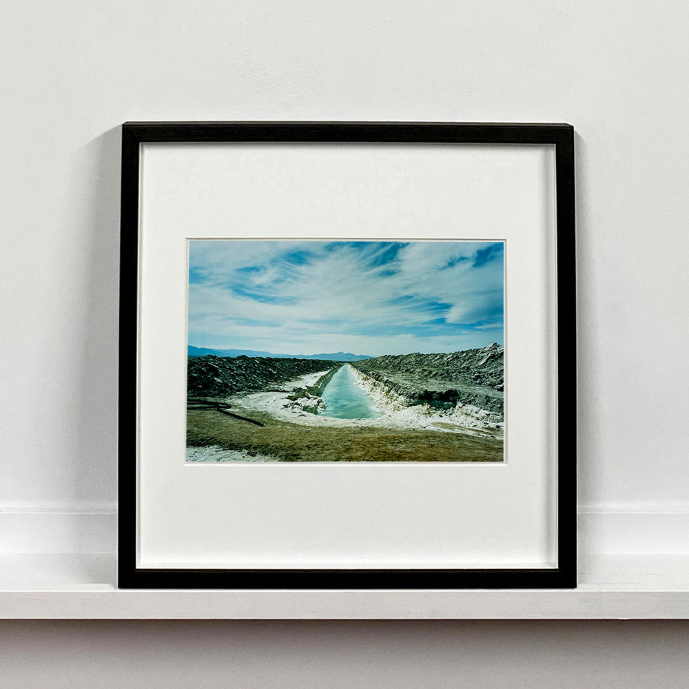 Black framed photograph by Richard Heeps. A watery white trench is dug into the centre going into the distance with grey mounds of earth either side. The sky is vast and blue, with light clouds.