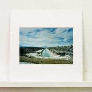 Mounted photograph by Richard Heeps. A watery white trench is dug into the centre going into the distance with grey mounds of earth either side. The sky is vast and blue, with light clouds.