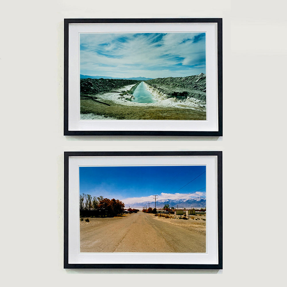 Two black framed photographs by Richard Heeps. The top photograph is of a watery white trench dug into the centre going into the distance with grey mounds of earth either side. The sky is vast and blue, with light clouds. The bottom photograph is a sandy dusty road going into the distance with bushy trees and small squarish buildings either side of the road. The sky is blue and there are mountains in the far distance.