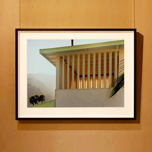 Black framed photograph by Richard Heeps.  The corner of the top of a cream colour building with big windows and vertical slats, on the left hand side are mountains bathing in the mist of dawn.