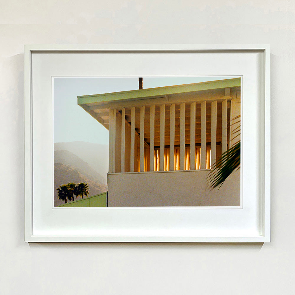 White framed photograph by Richard Heeps.  The corner of the top of a cream colour building with big windows and vertical slats, on the left hand side are mountains bathing in the mist of dawn.