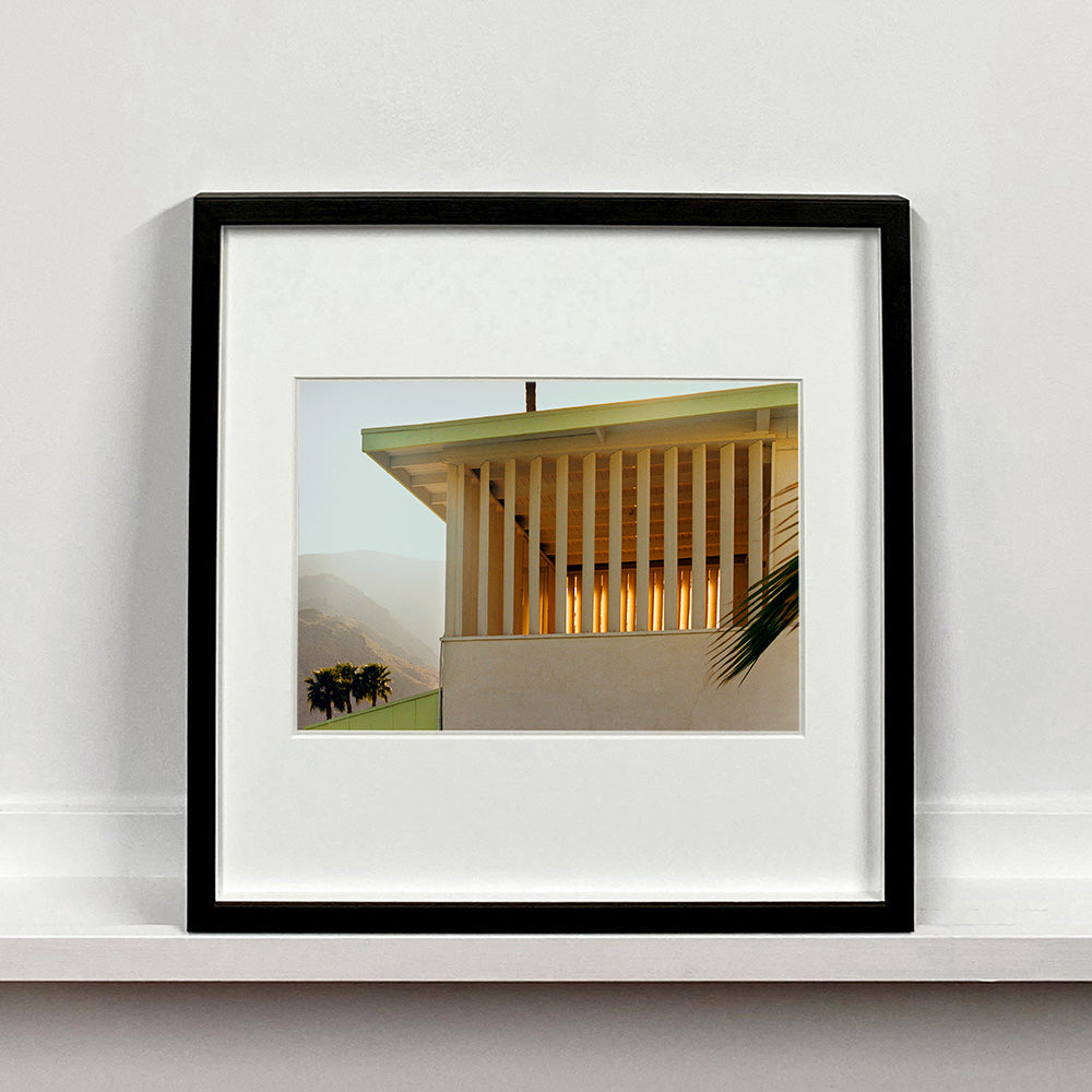 Black framed photograph by Richard Heeps.  The corner of the top of a cream colour building with big windows and vertical slats, on the left hand side are mountains bathing in the mist of dawn.