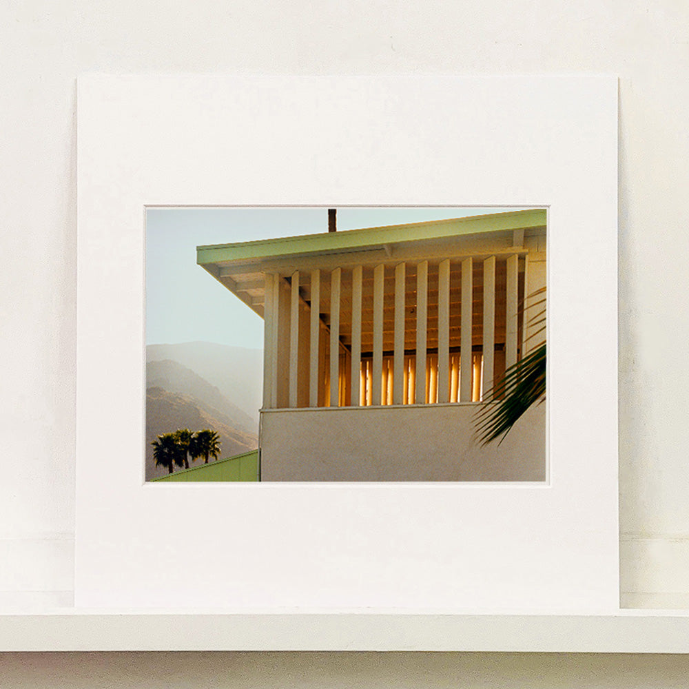 Mounted photograph by Richard Heeps.  The corner of the top of a cream colour building with big windows and vertical slats, on the left hand side are mountains bathing in the mist of dawn.