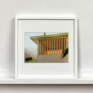 White framed photograph by Richard Heeps.  The corner of the top of a cream colour building with big windows and vertical slats, on the left hand side are mountains bathing in the mist of dawn.