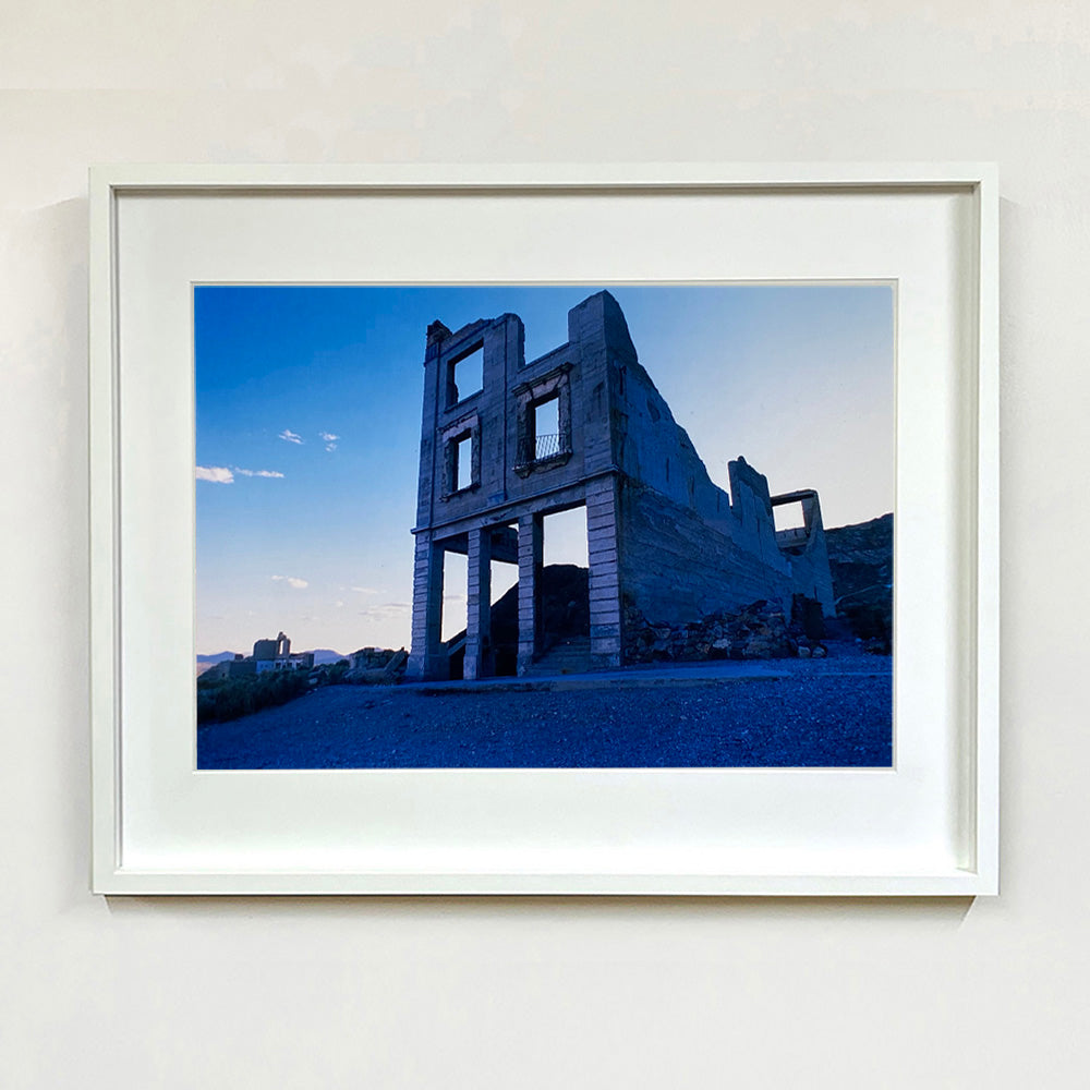 White framed photograph by Richard Heeps. The remnants of a rectangular building sits alone, surrounded by rubble and gravel in a blue light.