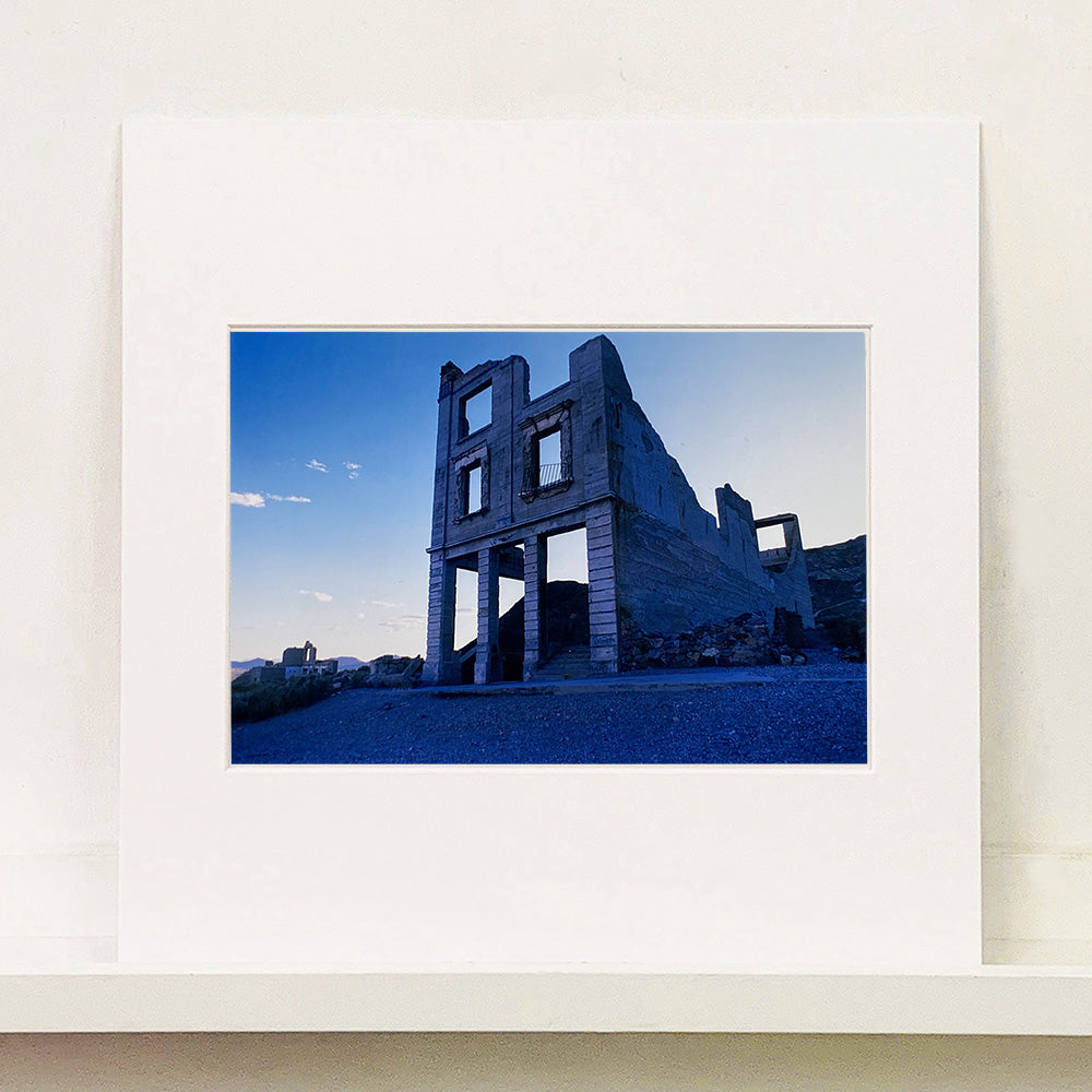 Mounted photograph by Richard Heeps. The remnants of a rectangular building sits alone, surrounded by rubble and gravel in a blue light.