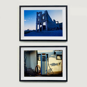 Two black framed photographs by Richard Heeps. The top photograph has the remnants of a rectangular building sitting alone, surrounded by rubble and gravel in a blue light. The bottom photograph has a retro Gas Pump white with blue sides, sitting outside the white slatted gas station.