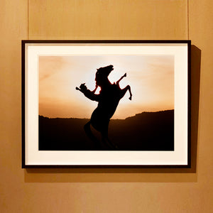 Photograph by Richard Heeps. The photograph shows a sunset and in the middle of the photograph is the silhouette of a statue of a cowboy on his bucking horse with one arm on the reins and the other held high.