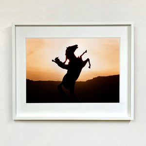 White framed photograph by Richard Heeps. The photograph shows a sunset and in the middle of the photograph is the silhouette of a statue of a cowboy on his bucking horse with one arm on the reins and the other held high.