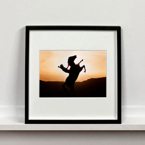 Black framed photograph by Richard Heeps. The photograph shows a sunset and in the middle of the photograph is the silhouette of a statue of a cowboy on his bucking horse with one arm on the reins and the other held high.