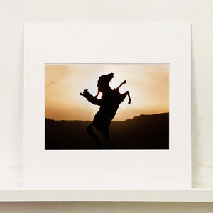 Mounted photograph by Richard Heeps. The photograph shows a sunset and in the middle of the photograph is the silhouette of a statue of a cowboy on his bucking horse with one arm on the reins and the other held high.