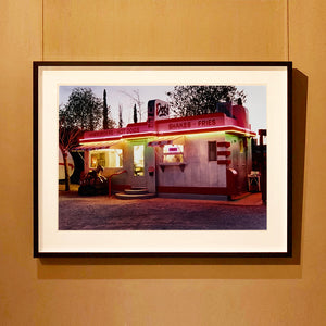Black framed photograph by Richard Heeps. This photograph depicts a one storey small building "Dot's Diner" brightly lit with a pink roof, with Hamburgers, Hot Dogs, Shakes, Fries written along the top width of the building.