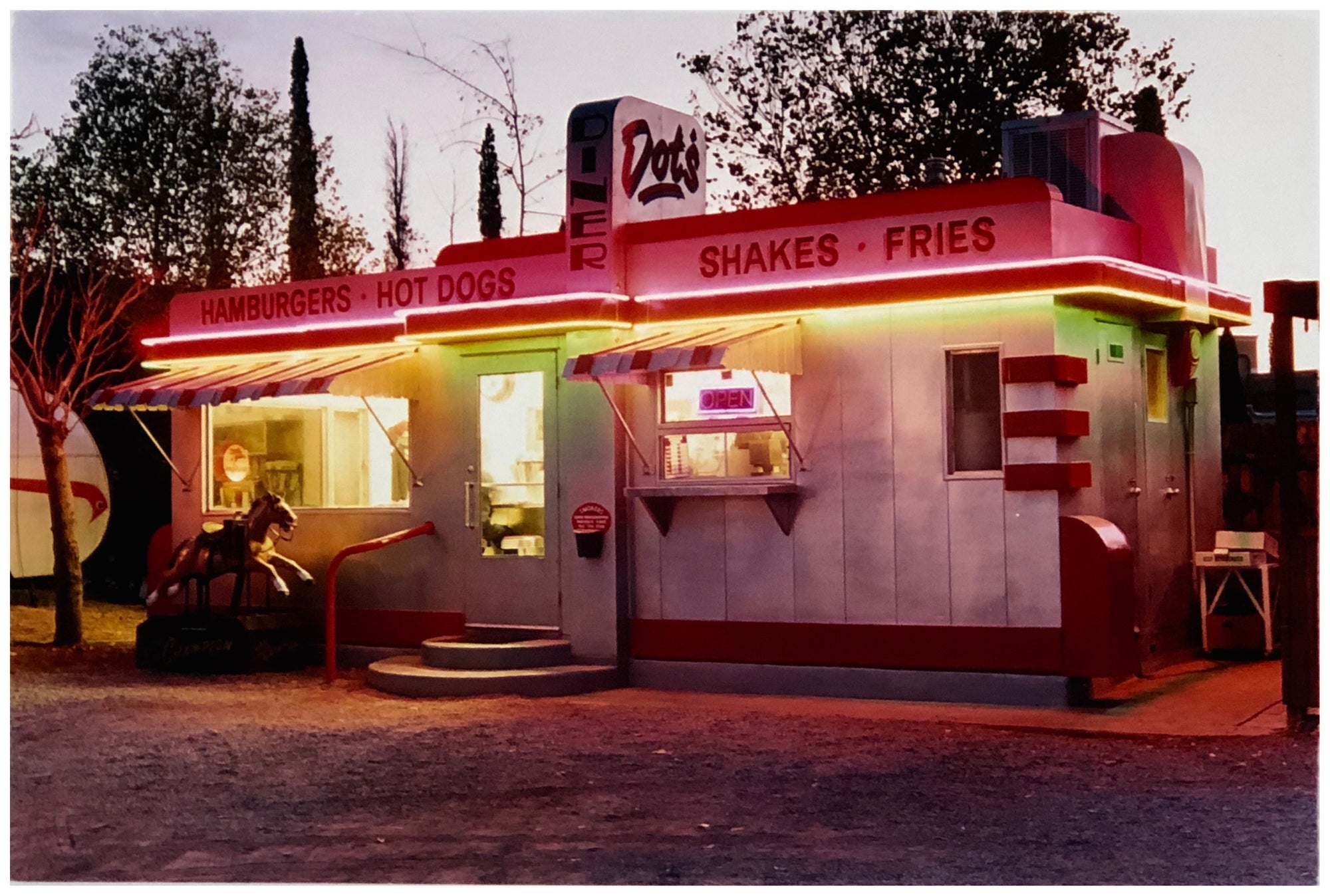 Photograph by Richard Heeps. This photograph depicts a one storey small building "Dot's Diner" brightly lit with a pink roof, with Hamburgers, Hot Dogs, Shakes, Fries written along the top width of the building.
