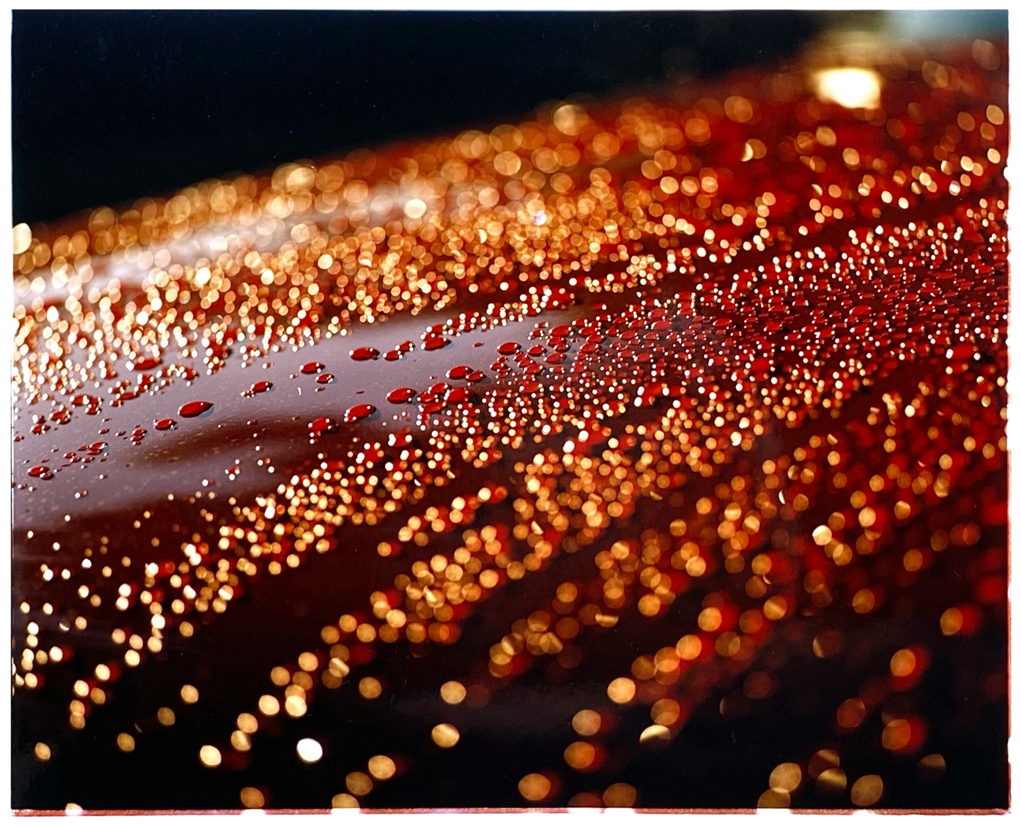 Photograph by Richard Heeps. The photograph is of a brown metal surface with water droplets on. The lighting makes the droplets appear in different colours of orange and brown.