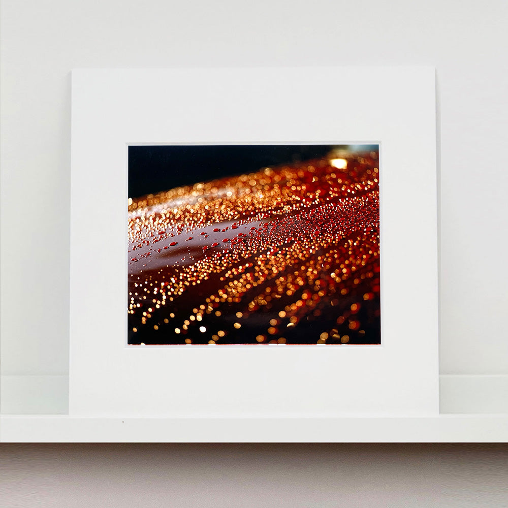 Mounted photograph by Richard Heeps. The photograph is of a brown metal surface with water droplets on. The lighting makes the droplets appear in different colours of orange and brown.