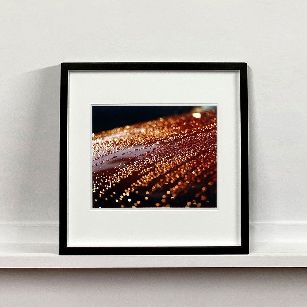 Black framed photograph by Richard Heeps. The photograph is of a brown metal surface with water droplets on. The lighting makes the droplets appear in different colours of orange and brown.