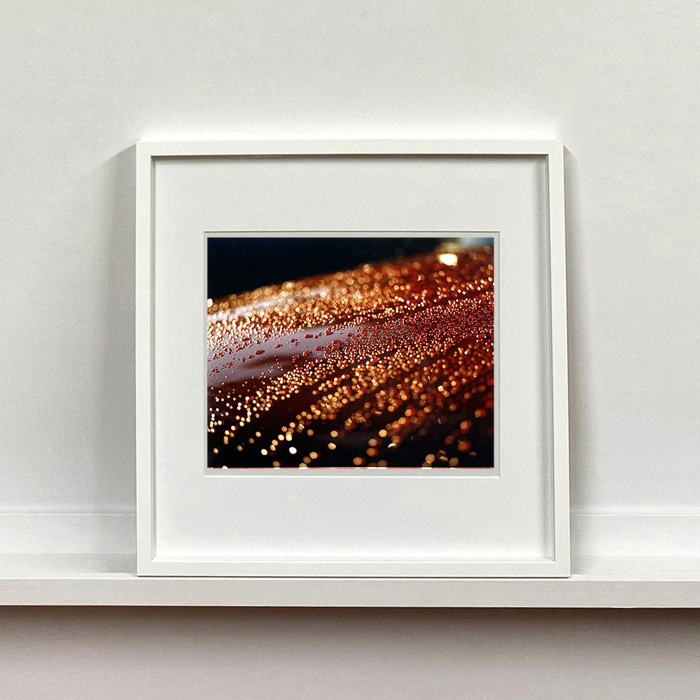 White framed photograph by Richard Heeps. The photograph is of a brown metal surface with water droplets on. The lighting makes the droplets appear in different colours of orange and brown.