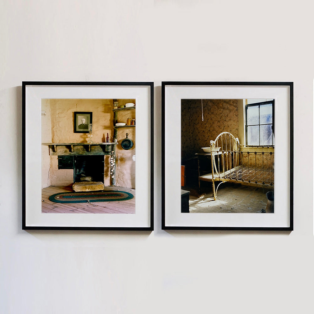 Black framed photographs by Richard Heeps. The first is from the Film set of 'The Outlaw Josey Wales' featuring a wooden fireplace and a black and white photo over the mantlepiece. The second photograph is of a bedroom in an abandoned town featuring a bed frame, stand and wash basin.