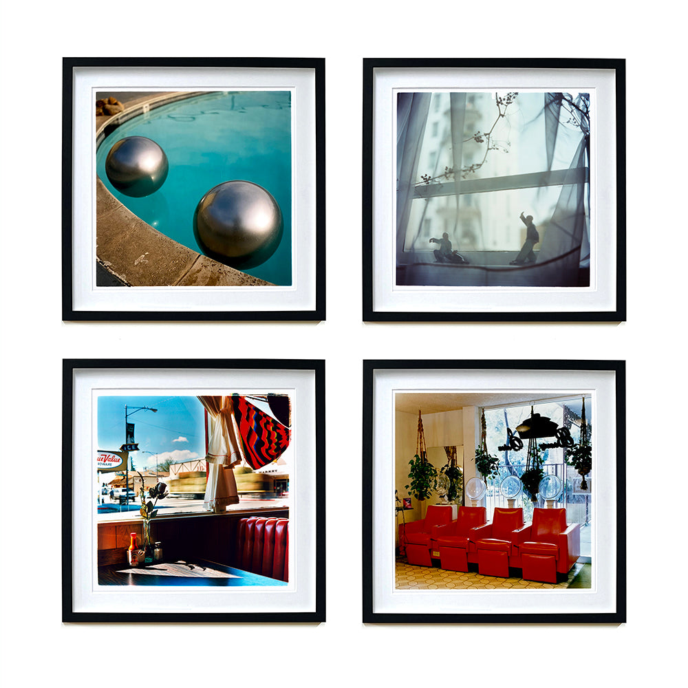 4 Black framed photographs by Richard Heeps. The top left hand photograph is of the corner of a circular swimming pool with two metallic silver beach balls floating on the water. The top right photo is ceramic figures sitting on a windowsill. The bottom left hand photo is a booth in an American diner, the bottom right is red chairs lined up in a barbers.