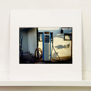 Mounted photograph by Richard Heeps. A vintage petrol pump with a white front and blue sides, sitting outside a white slatted building.