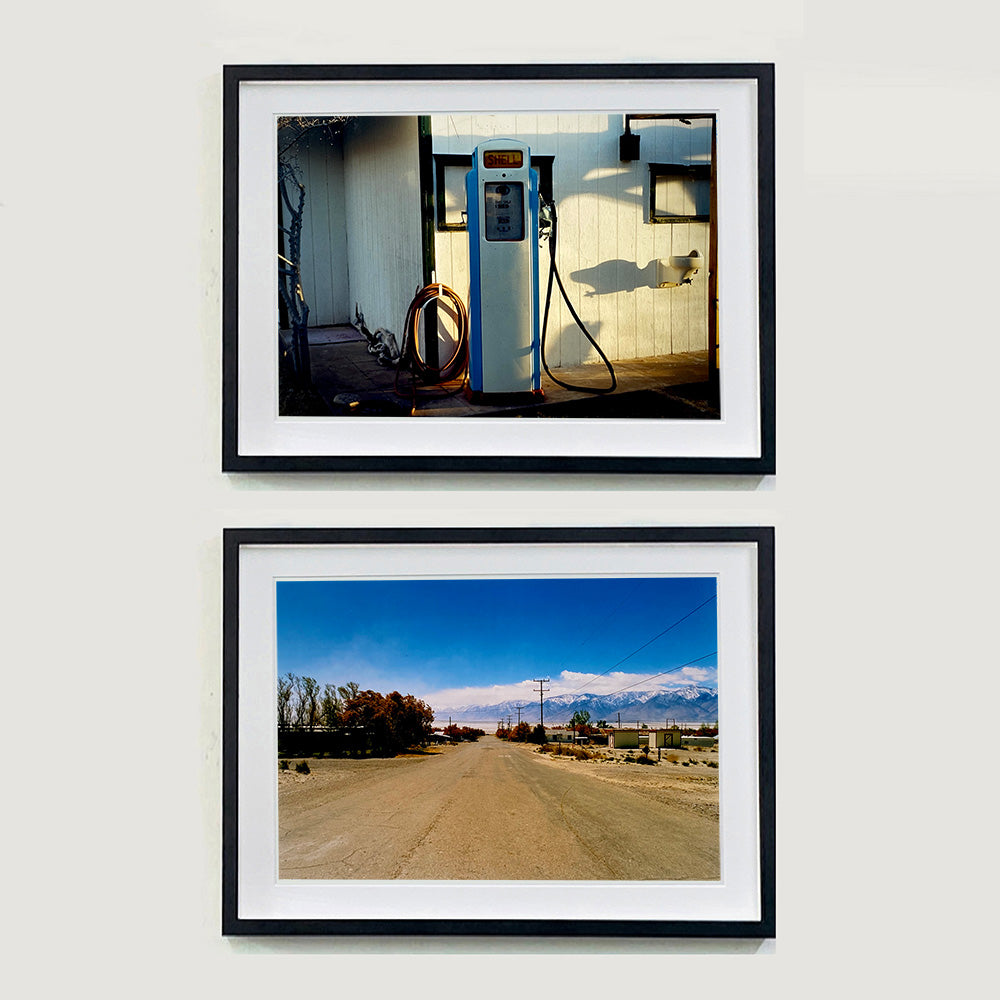 Two black framed photographs held by Richard Heeps. The top photograph depicts a vintage petrol pump with a white front and blue sides, sitting outside a white slatted building.  The bottom photograph shows a dusty road in the middle, heading towards the snow capped mountains in the distance, on the right are brown bushes and trees and on the left, single level concrete buildings. 