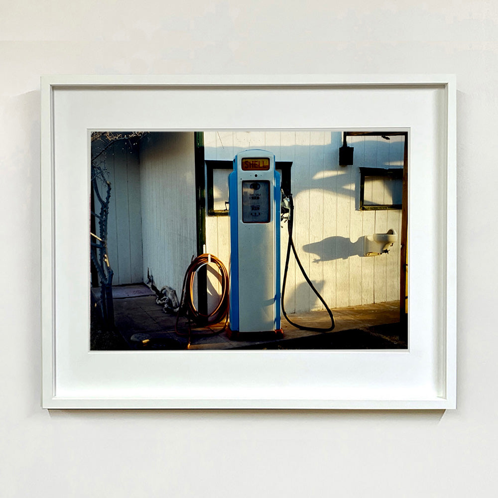 White framed photograph by Richard Heeps. A vintage petrol pump with a white front and blue sides, sitting outside a white slatted building.