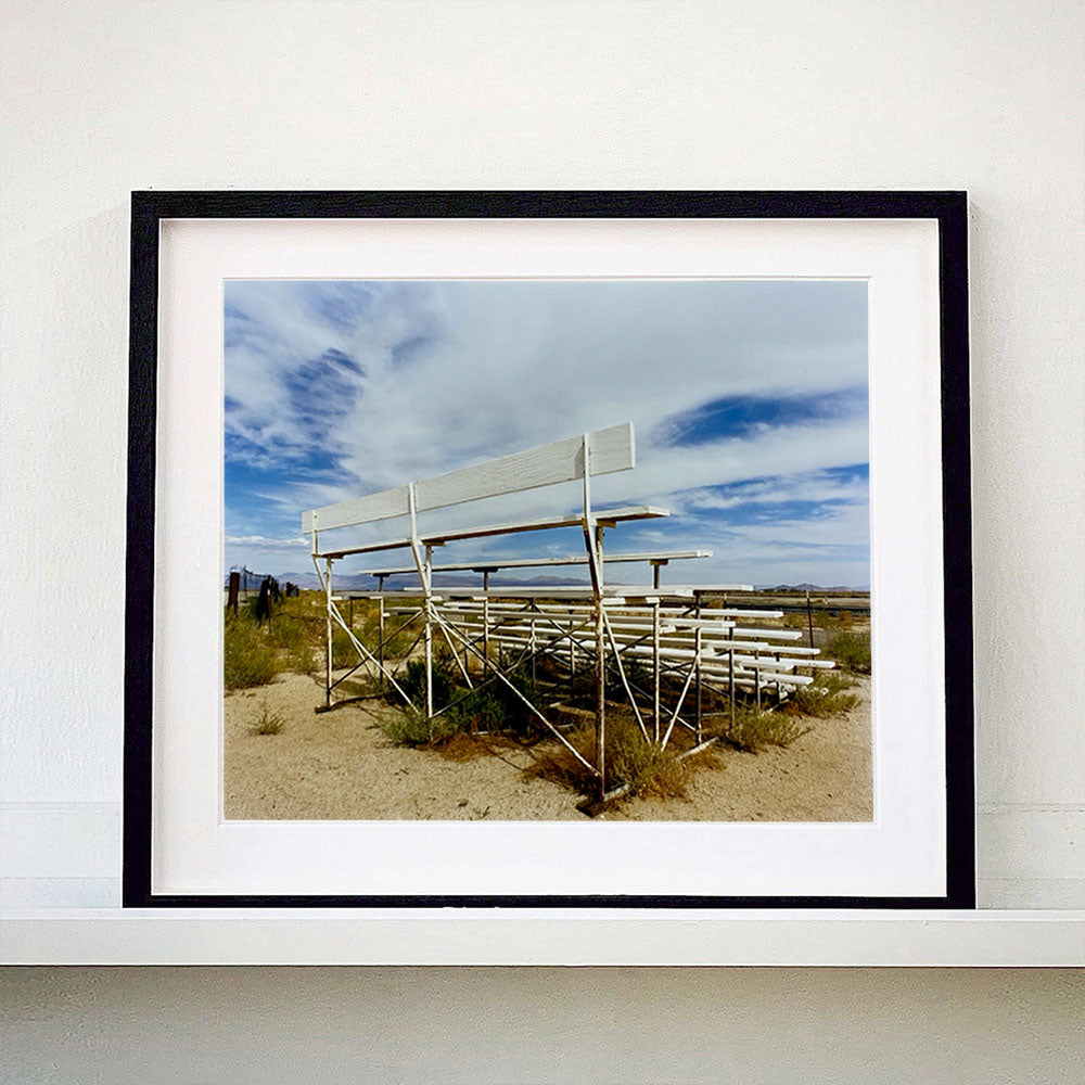 Black framed photograph by Richard Heeps. The back view of a basic white painted grandstand sits in the middle of this photograph. Grass grows up from its base and it sits alone on a sandy ground.