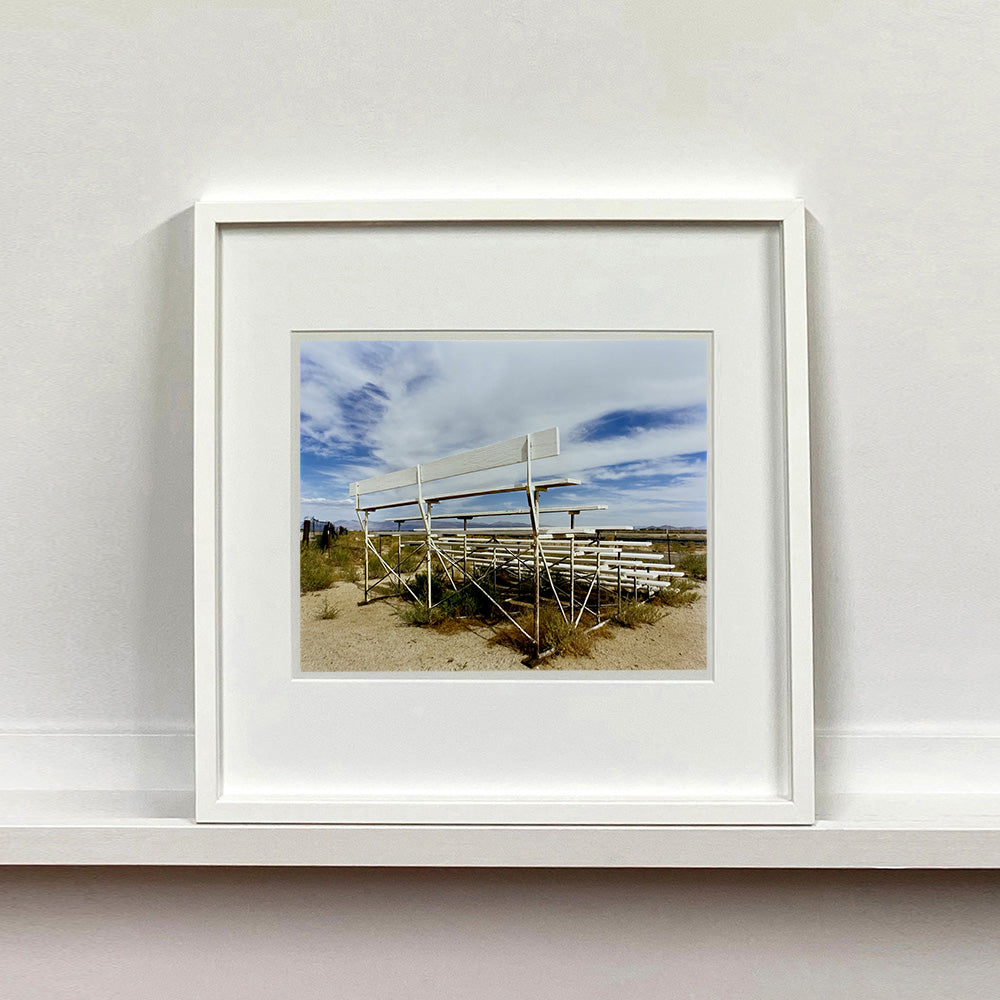 White framed photograph by Richard Heeps. The back view of a basic white painted grandstand sits in the middle of this photograph. Grass grows up from its base and it sits alone on a sandy ground.