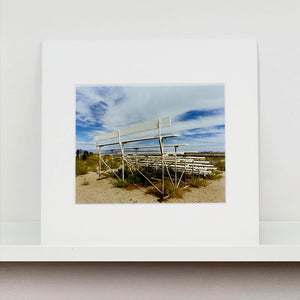 Mounted photograph by Richard Heeps. The back view of a basic white painted grandstand sits in the middle of this photograph. Grass grows up from its base and it sits alone on a sandy ground.