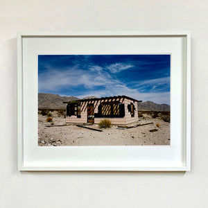 White framed photograph by Richard Heeps. An abandoned building sitting alone in a desert, tumble weed on the ground and hills in the background. A blue sky with white clouds.