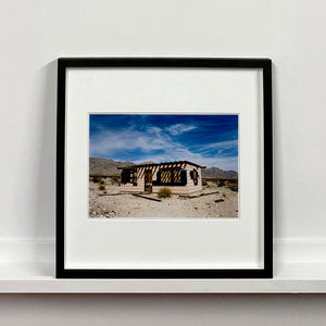 Black framed photograph by Richard Heeps. An abandoned building sitting alone in a desert, tumble weed on the ground and hills in the background. A blue sky with white clouds.