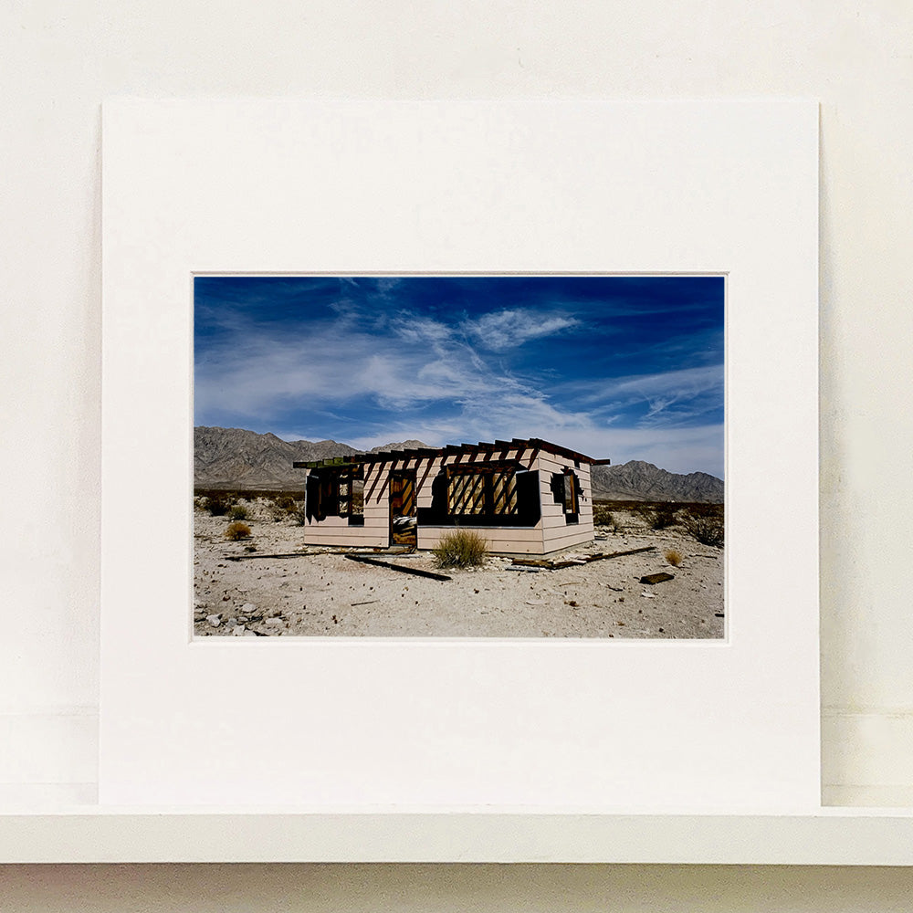 Mounted photograph by Richard Heeps. An abandoned building sitting alone in a desert, tumble weed on the ground and hills in the background. A blue sky with white clouds.