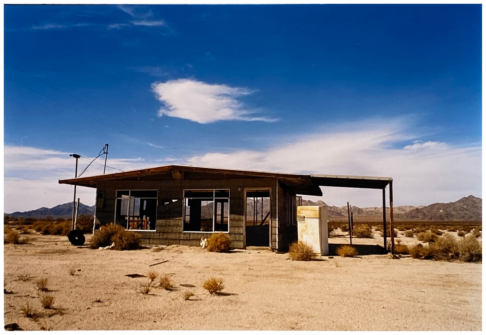 Photograph by Richard Heeps. An abandoned building sitting alone in a desert, tumble weed on the ground and hills in the background. A blue sky with white clouds.