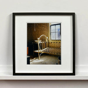 Black framed photograph by Richard Heeps. A run down now unused room with the metal surround of a cot bed and no mattress, at the end of the bed a wash stand with a bowl on top. Light shines in the room from the window.