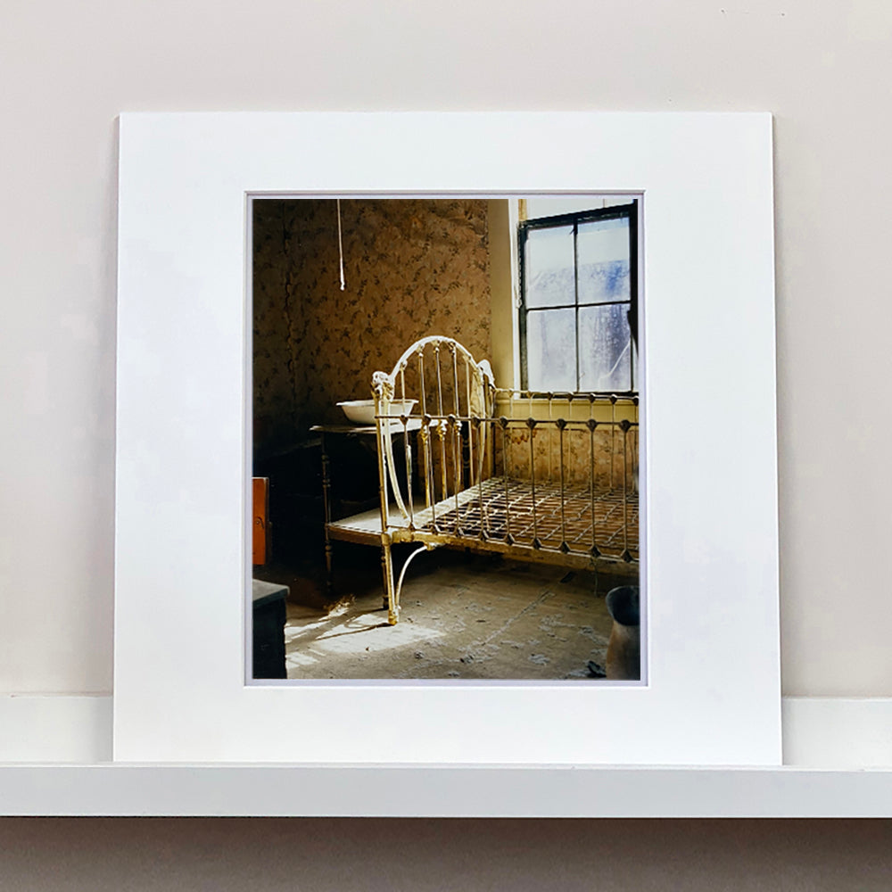 Mounted photograph by Richard Heeps. A run down now unused room with the metal surround of a cot bed and no mattress, at the end of the bed a wash stand with a bowl on top. Light shines in the room from the window.
