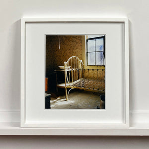 White framed photograph by Richard Heeps. A run down now unused room with the metal surround of a cot bed and no mattress, at the end of the bed a wash stand with a bowl on top. Light shines in the room from the window.