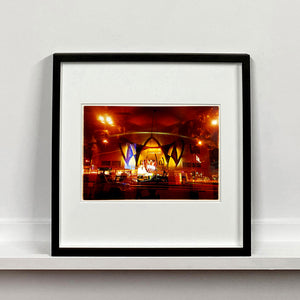 Black framed photograph by Richard Heeps. Photograph taken at night of the front of a brightly lit hotel. The scene is bathed in golden and reddish brown colours. The name of the hotel is La Concha and its name is written in white neon against a red and golden shell design. The photograph is taken through glass and there are reflections of cars as well as the room from which the photograph is taken.