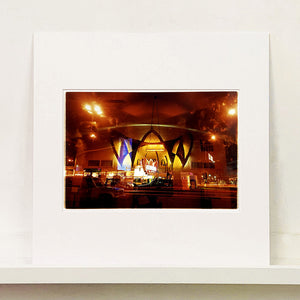 Mounted photograph by Richard Heeps. Photograph taken at night of the front of a brightly lit hotel. The scene is bathed in golden and reddish brown colours. The name of the hotel is La Concha and its name is written in white neon against a red and golden shell design. The photograph is taken through glass and there are reflections of cars as well as the room from which the photograph is taken.