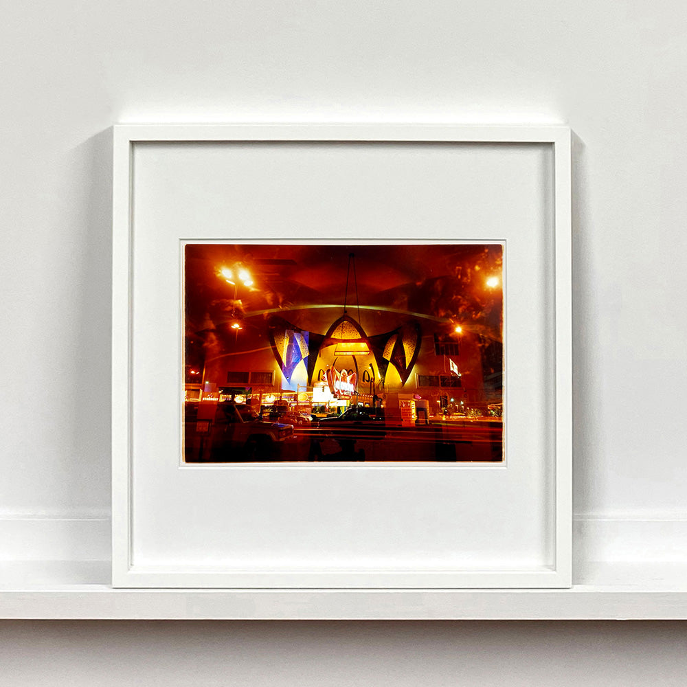White framed photograph by Richard Heeps. Photograph taken at night of the front of a brightly lit hotel. The scene is bathed in golden and reddish brown colours. The name of the hotel is La Concha and its name is written in white neon against a red and golden shell design. The photograph is taken through glass and there are reflections of cars as well as the room from which the photograph is taken.