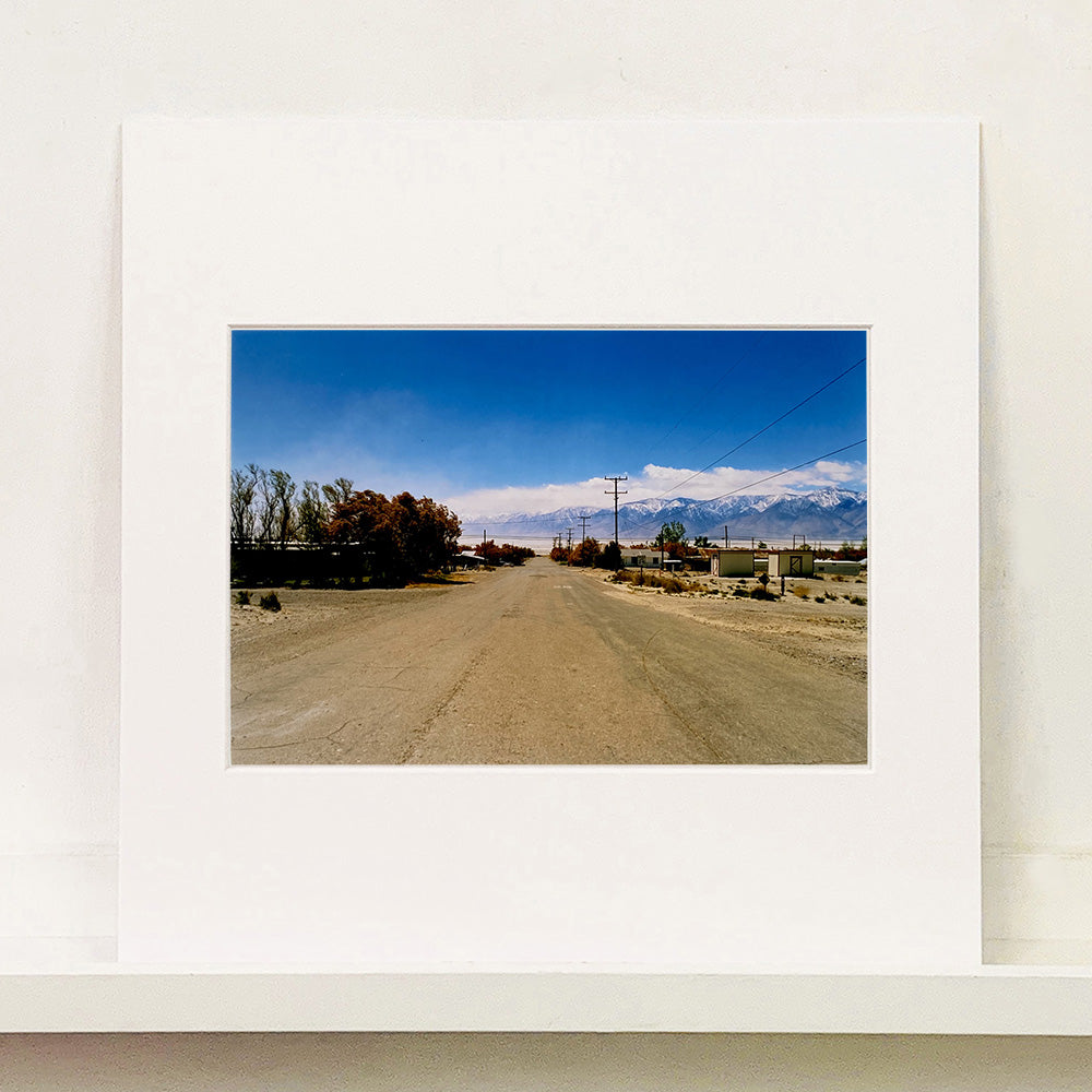 Photograph by Richard Heeps. A dusty road in the middle, heading towards the snow capped mountains in the distance, on the right are brown bushes and trees and on the left, single level concrete buildings.